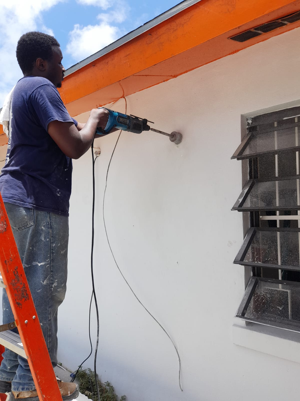 Technician on a ladder outside, core-drilling through an exterior wall to run an AC line set