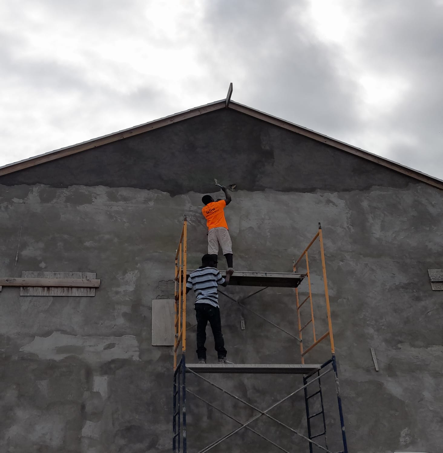 Two workers on scaffolding troweling fresh plaster onto a tall exterior gable wall