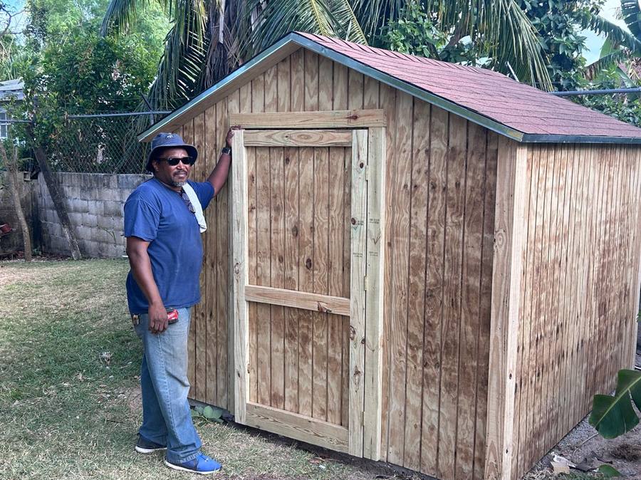 Finished custom shed in natural wood with a red shingle roof, owner standing beside it